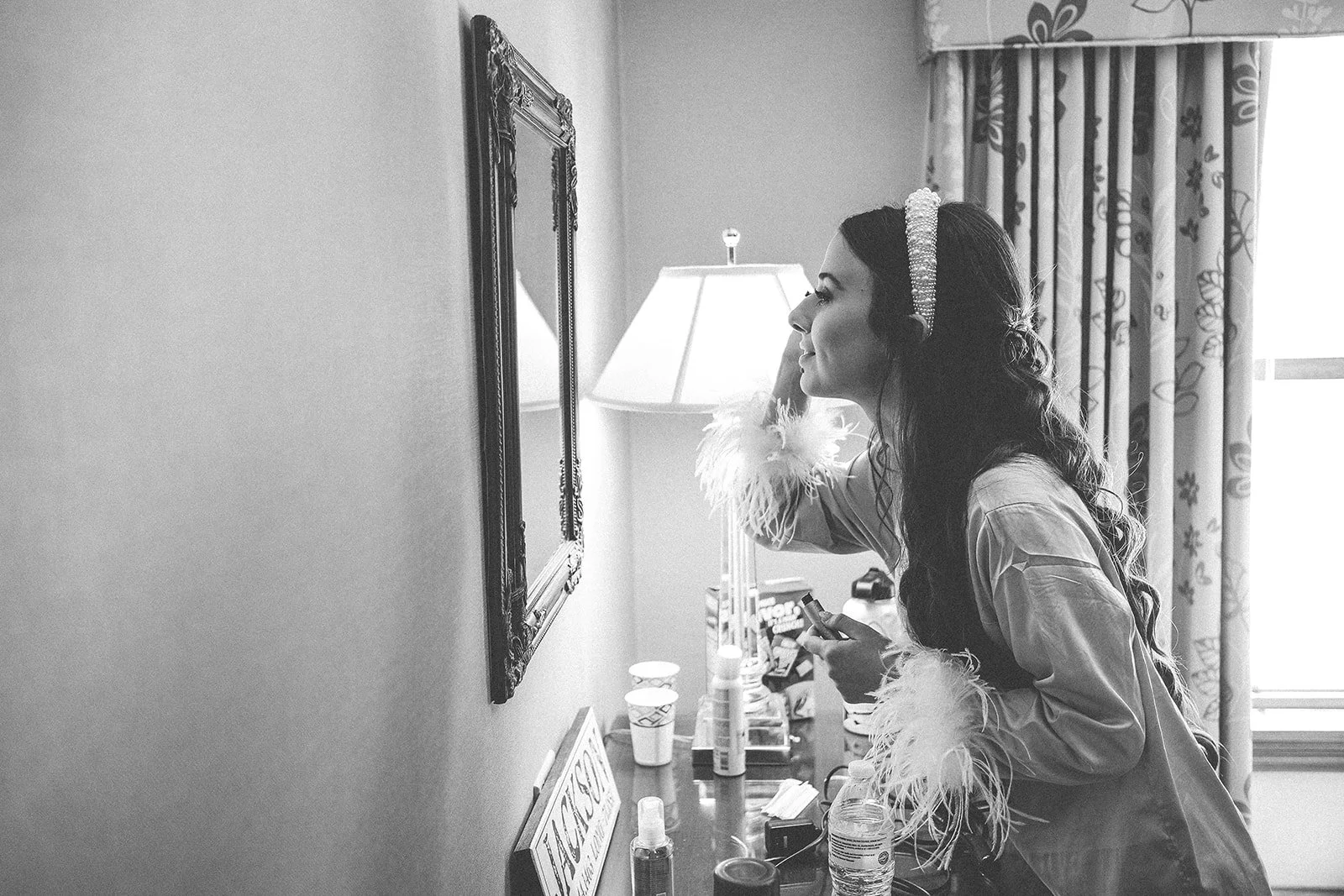 A woman with dark hair wearing a tiara and a robe with feather trim is looking into a mirror in a hotel room, applying makeup or grooming. The room has a side table with various toiletries, snacks, and a water bottle, and curtains with a floral patte