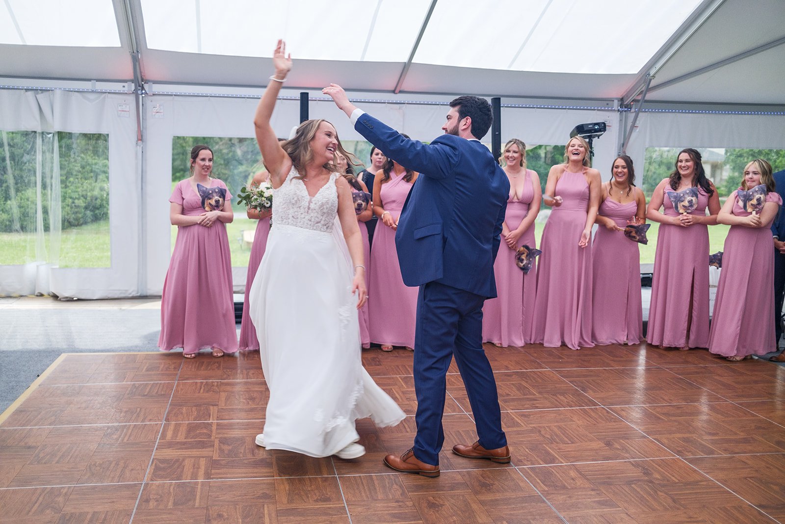 A bride and groom dance together at their wedding reception, surrounded by bridesmaids in pink dresses under a white tent.