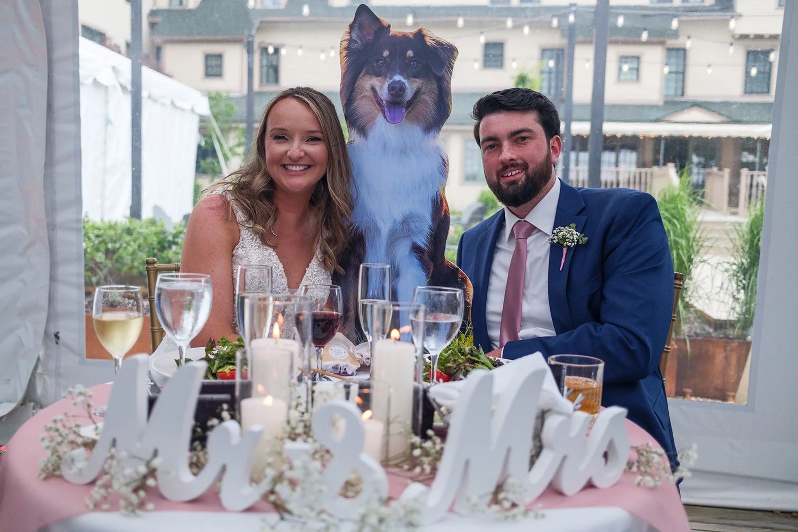A bride and groom sitting at a table during their wedding celebration, with a cardboard cutout of a dog behind them. The table is decorated with candles, flowers, and drink glasses.