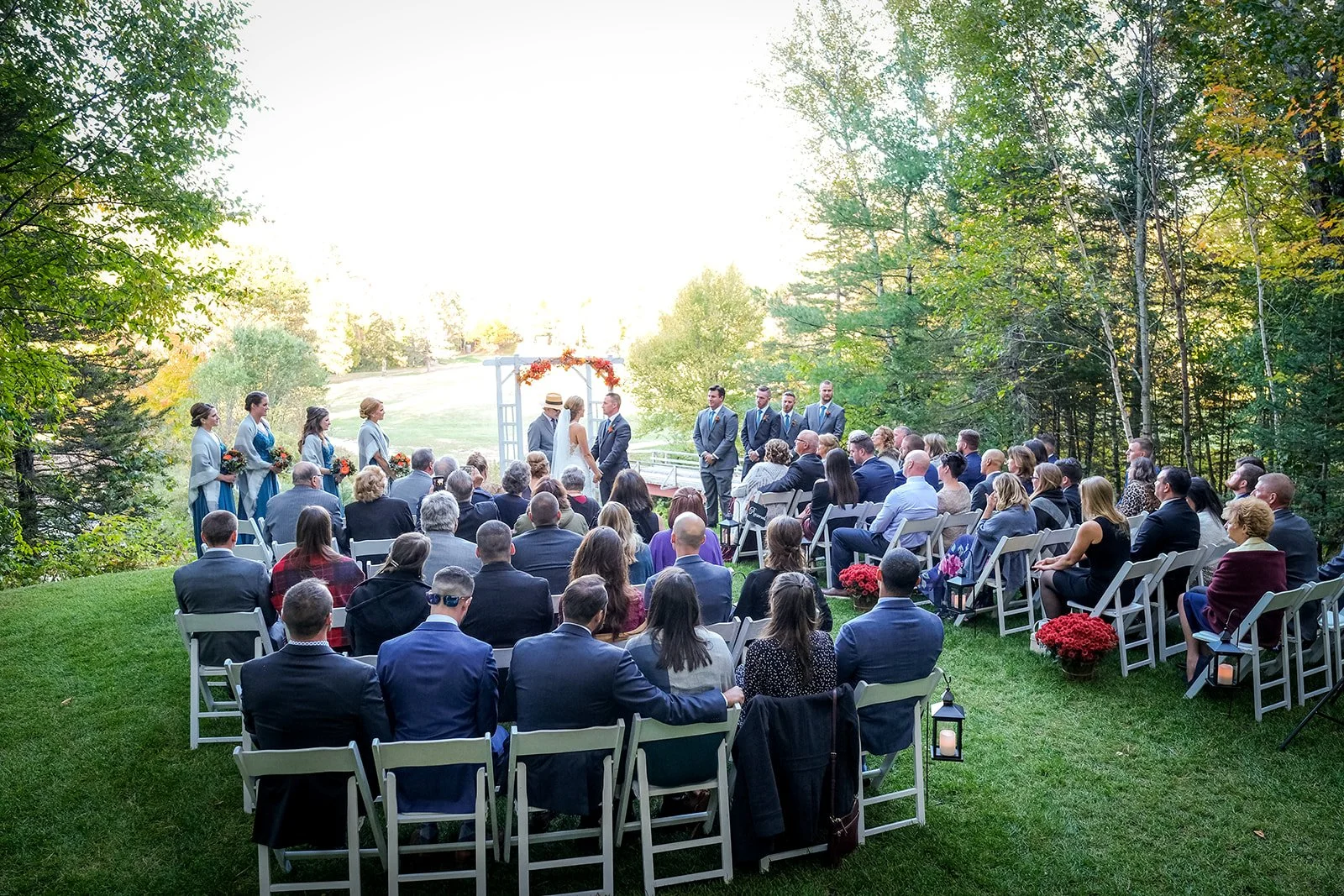 Outdoor wedding ceremony with bride and groom standing before officiant, surrounded by witnesses and guests seated on chairs, in a green, wooded setting.