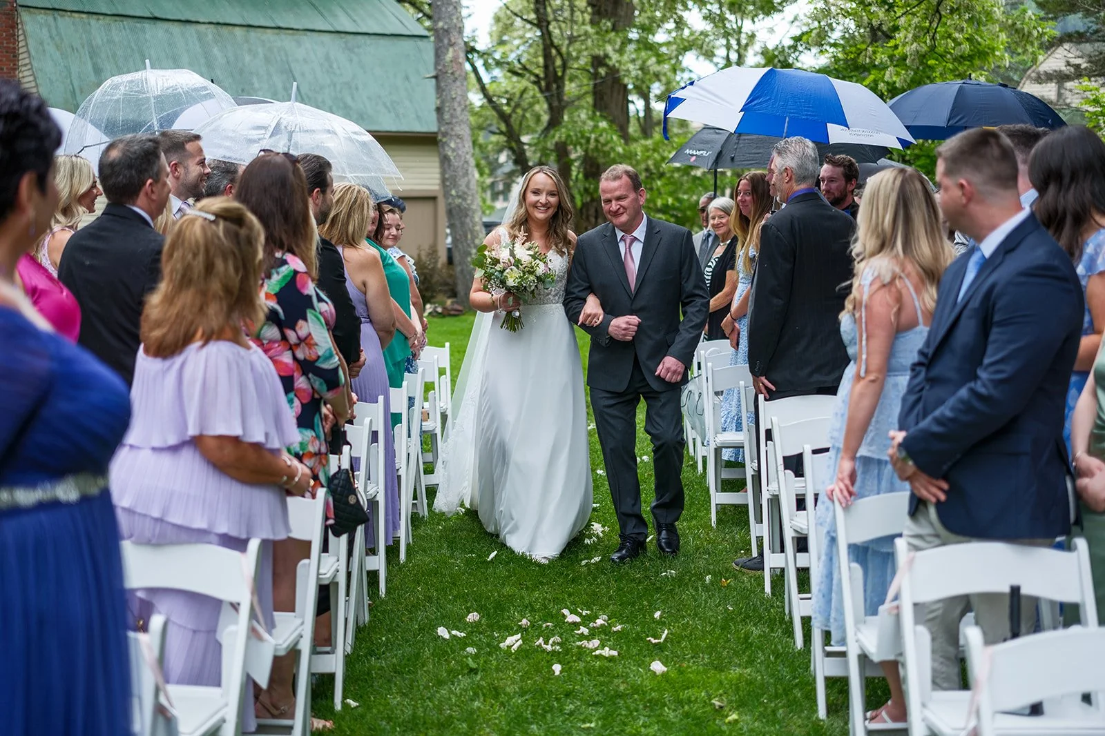 Bride walking down the aisle arm-in-arm with her father at an outdoor wedding ceremony, with guests sitting and standing under umbrellas due to rain.