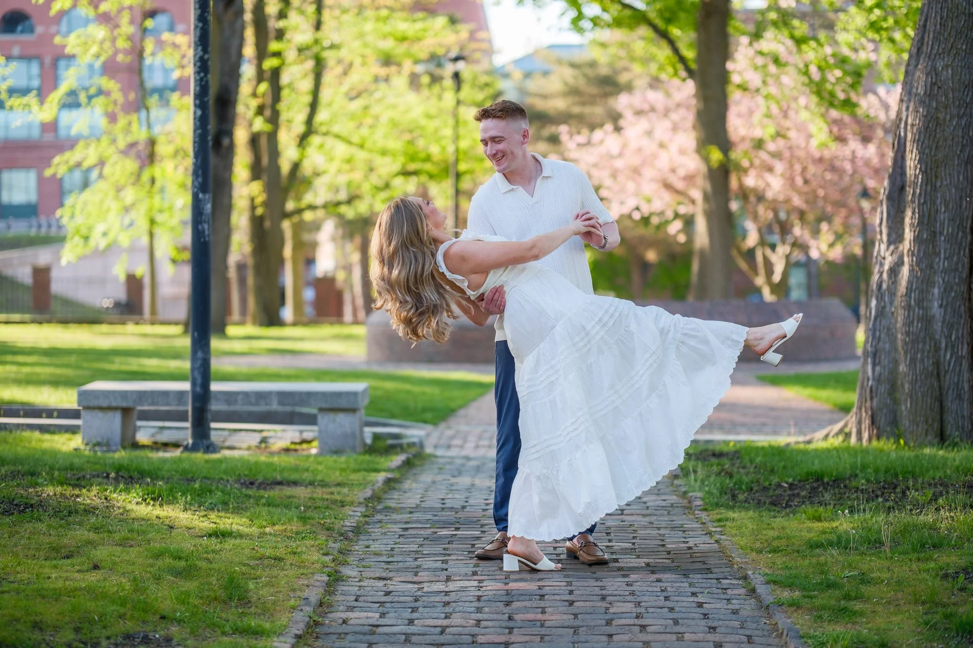 Melanie leans back into Connor's arms on a white garden bench beneath blooming tree branches during their Prescott Park engagement session in Portsmouth, NH