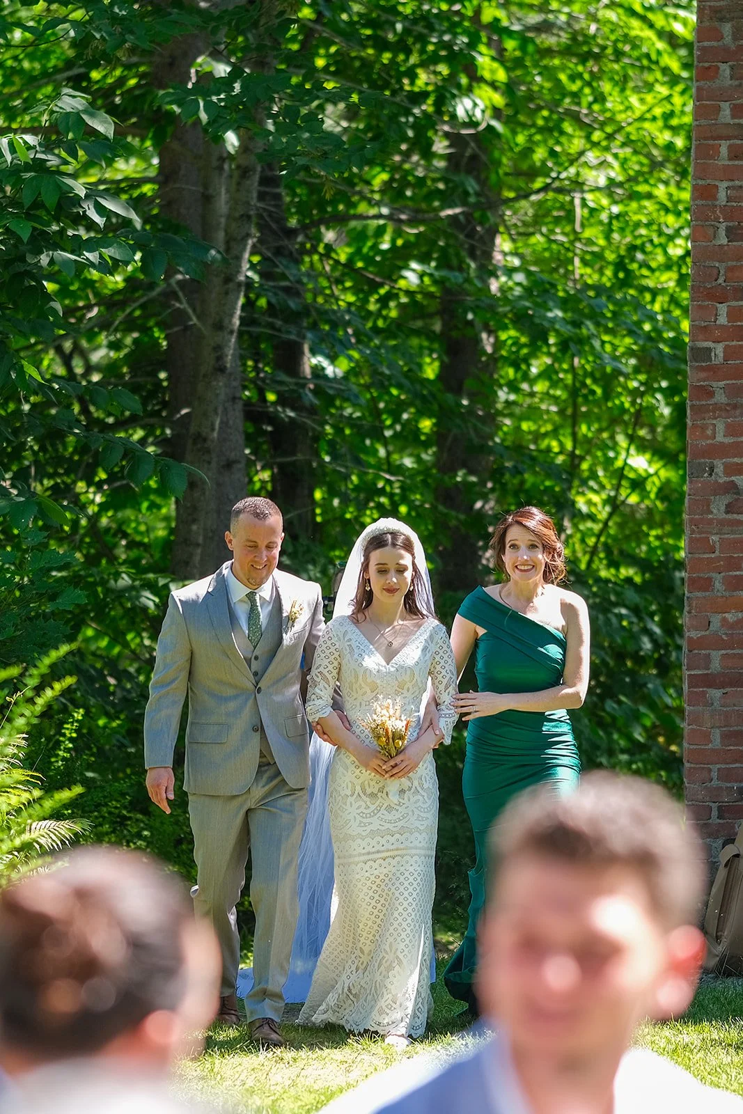 A bride in a white lace wedding dress walking outdoors with a man in a light-colored suit and a woman in a green dress beside her, surrounded by greenery and trees.