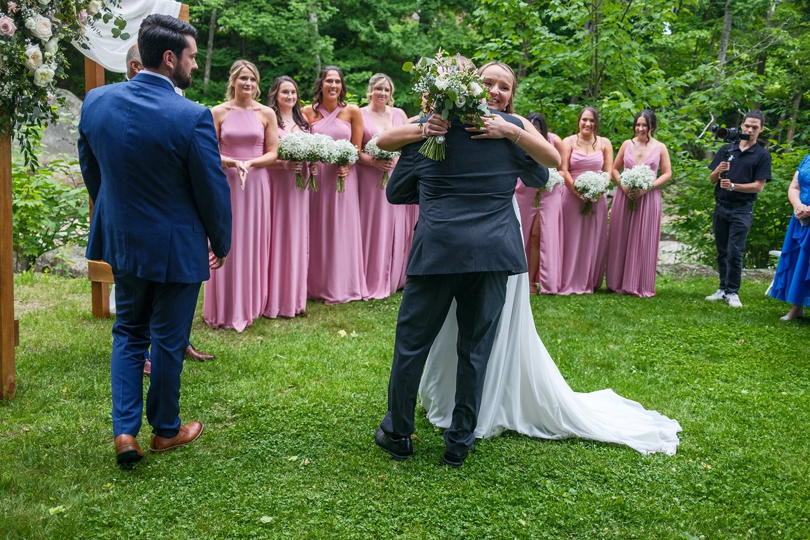 A wedding ceremony outdoors in a green forest area. The bride and groom are embracing, with the bride smiling and holding a bouquet of flowers. Bridesmaids dressed in pink gowns stand behind them, holding bouquets, while a man in black with a camera 