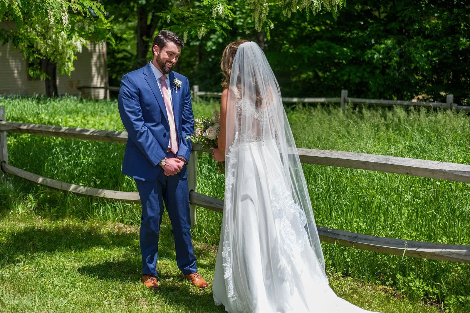 A bride and groom exchanging vows outdoors on a sunny day, with the groom in a blue suit and the bride in a white wedding dress and veil, standing near a wooden fence with green trees in the background.