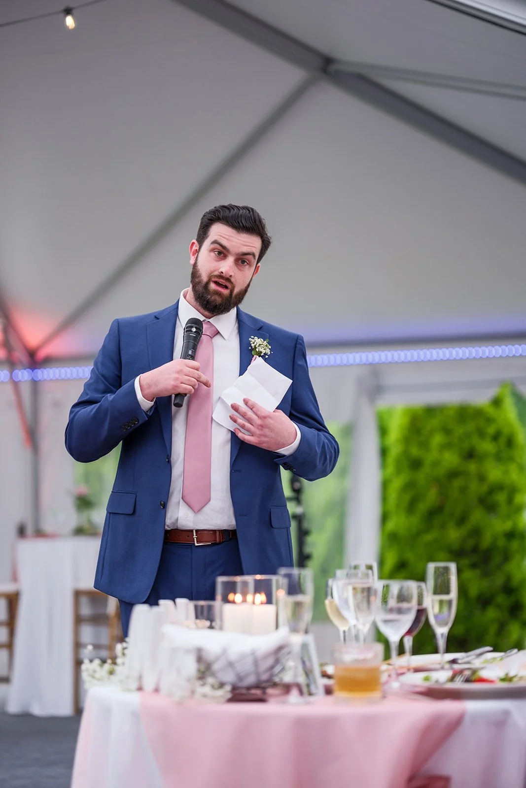 A man in a blue suit and pink tie giving a speech at a wedding reception under a tent, holding a microphone and a piece of paper, with a decorated table in front of him.