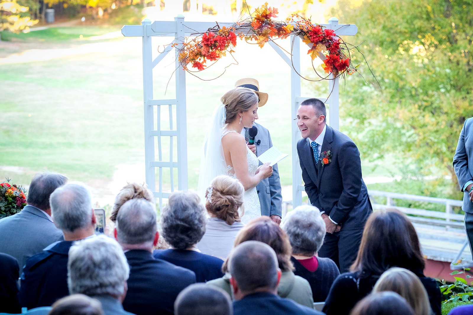 A wedding ceremony outdoors with a bride and groom exchanging vows under an arch decorated with fall-colored leaves and flowers, surrounded by seated guests.
