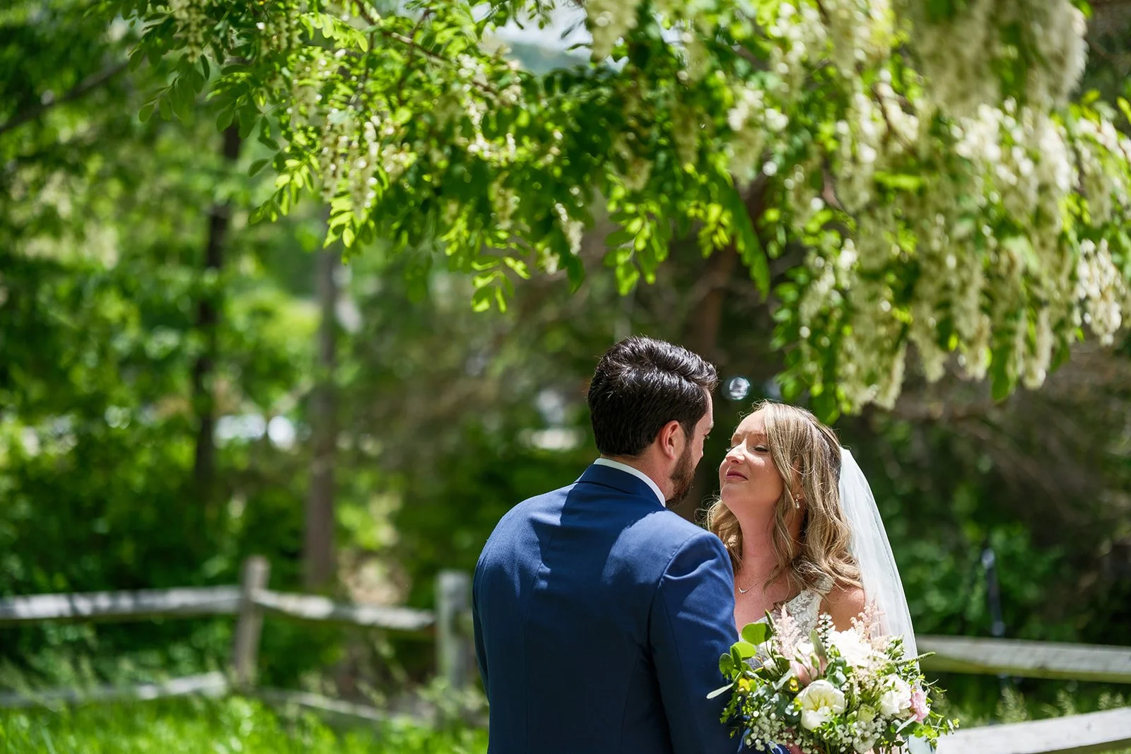 A bride and groom standing close together outdoors, with the bride holding a bouquet of white flowers, under green trees in a garden setting.