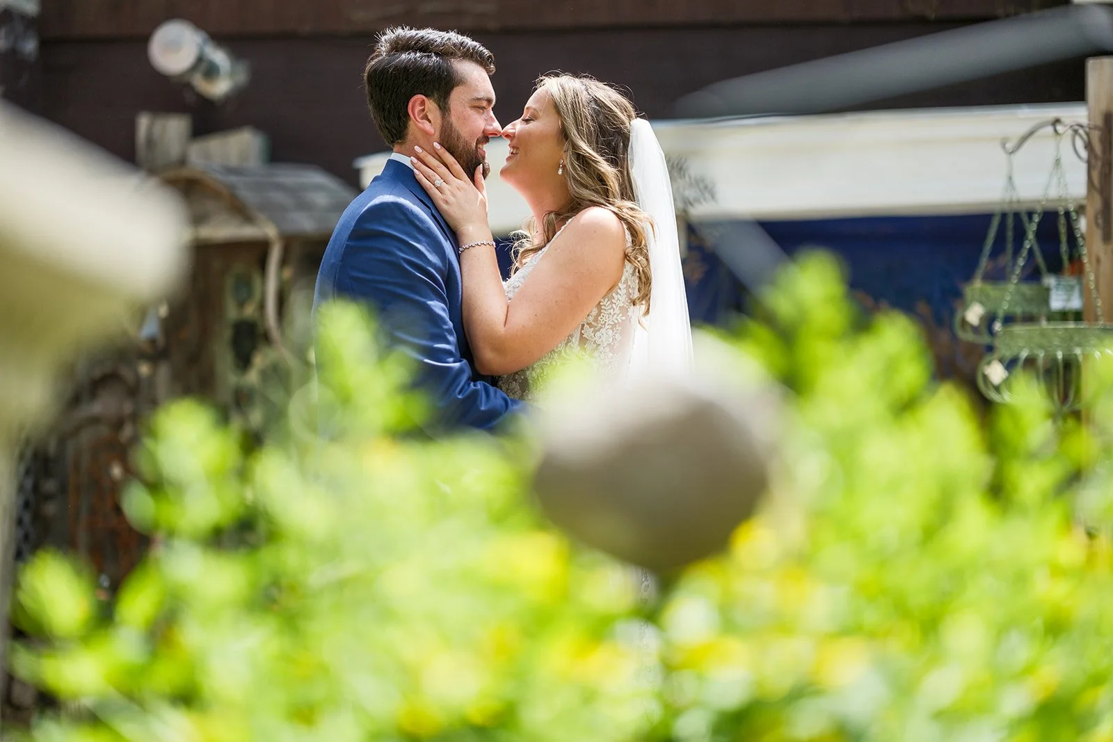 A bride and groom share a kiss during their wedding, with the bride gently holding the groom's face, both dressed in wedding attire, surrounded by blurred greenery and outdoor setting.