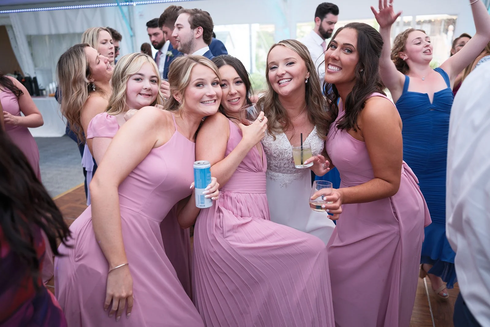 A group of women in pink and purple dresses at a celebration or wedding, smiling and holding drinks, dancing and enjoying themselves under a large white tent.