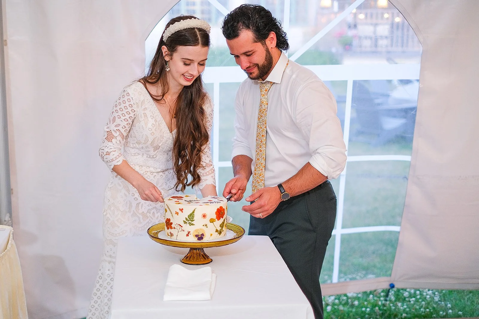 A man and woman are cutting a cake together at a celebration event, with the woman wearing a white dress and the man wearing a white shirt and patterned tie.