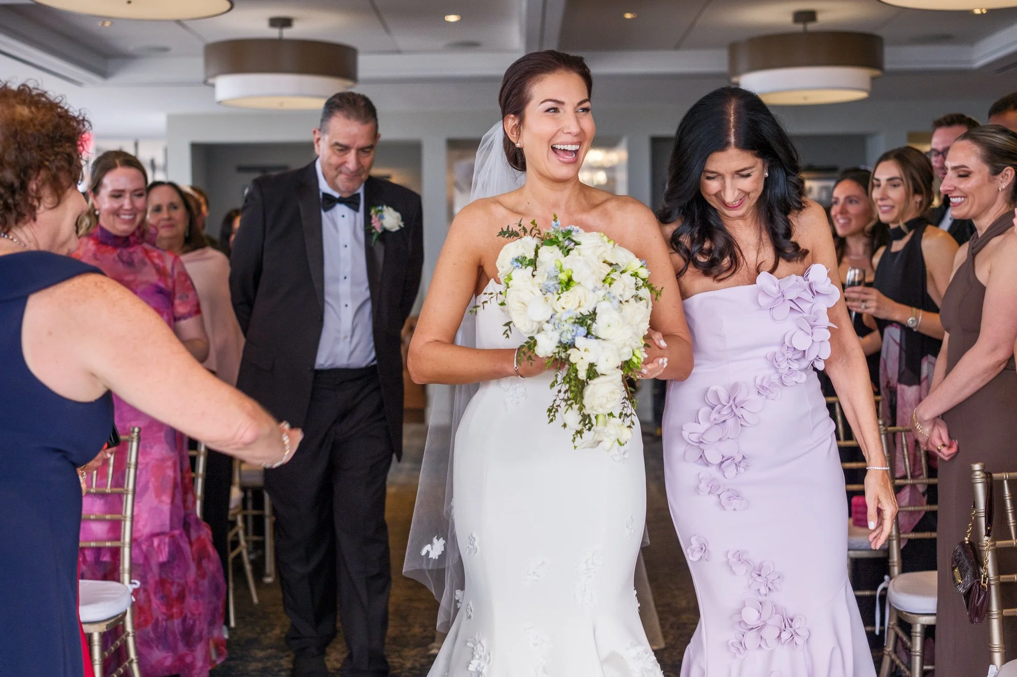 Bride beaming and laughing as she walks down the aisle with her parents during the UMass Club Boston Jewish wedding ceremony