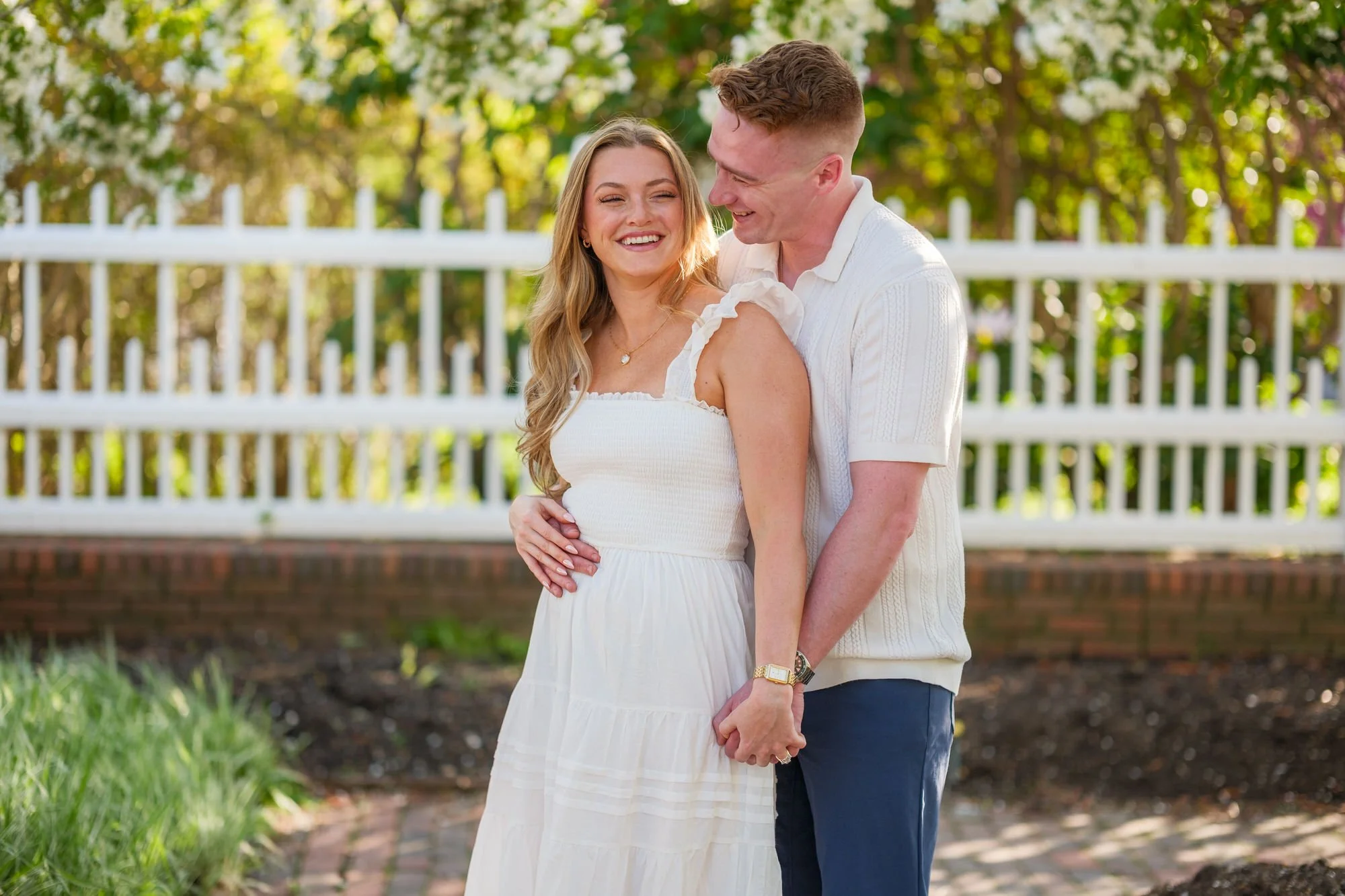 Melanie and Connor sit together on a brick garden wall beneath cascading white flowering tree branches at Prescott Park, Portsmouth, NH