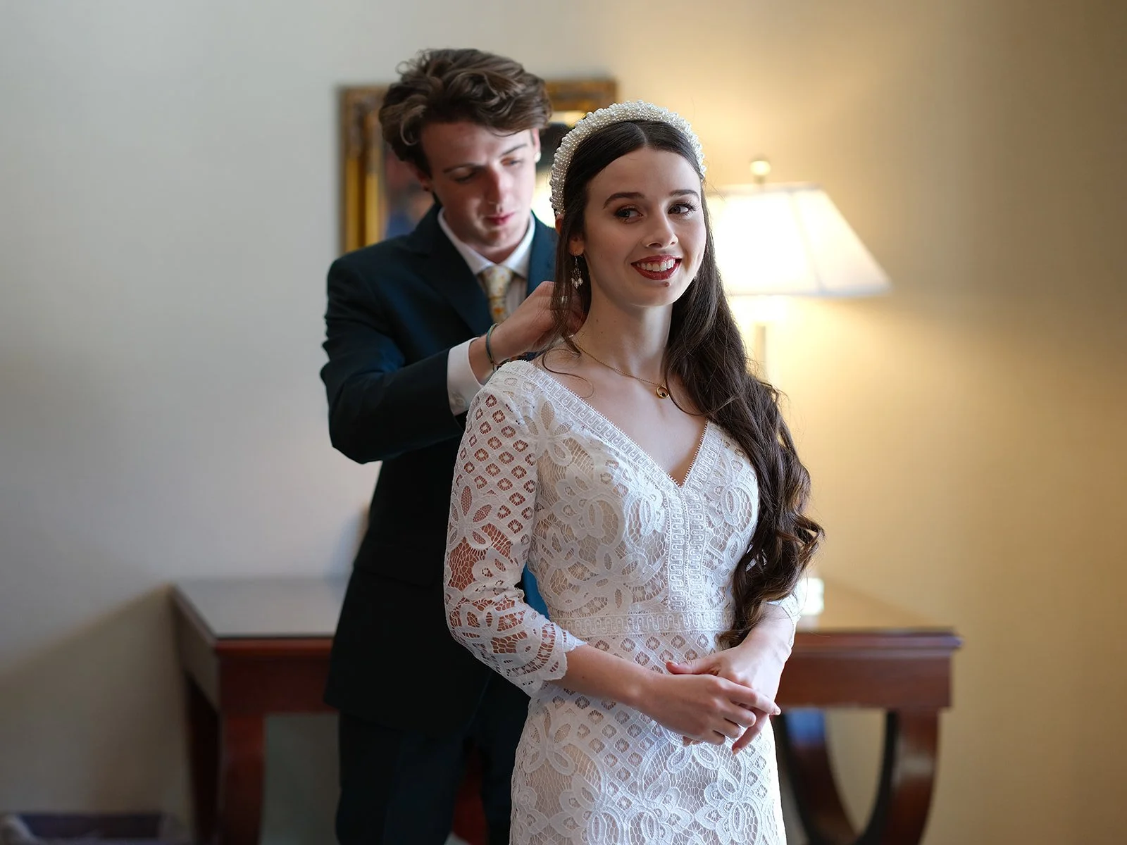 A woman in a white lace wedding dress is smiling as a man in a suit adjusts her hair.