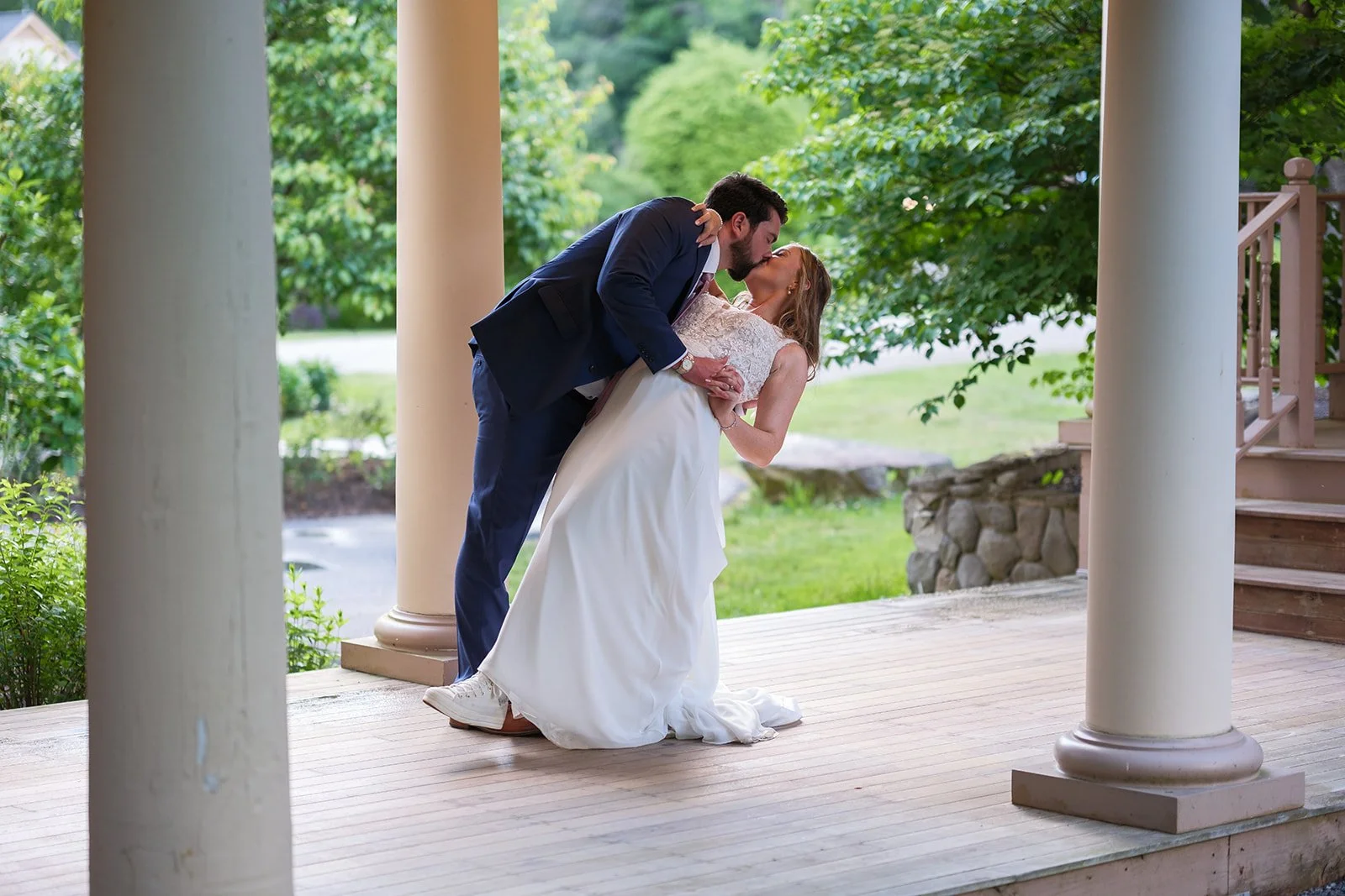 A groom dips and kisses a bride on a porch with large white columns, surrounded by greenery.