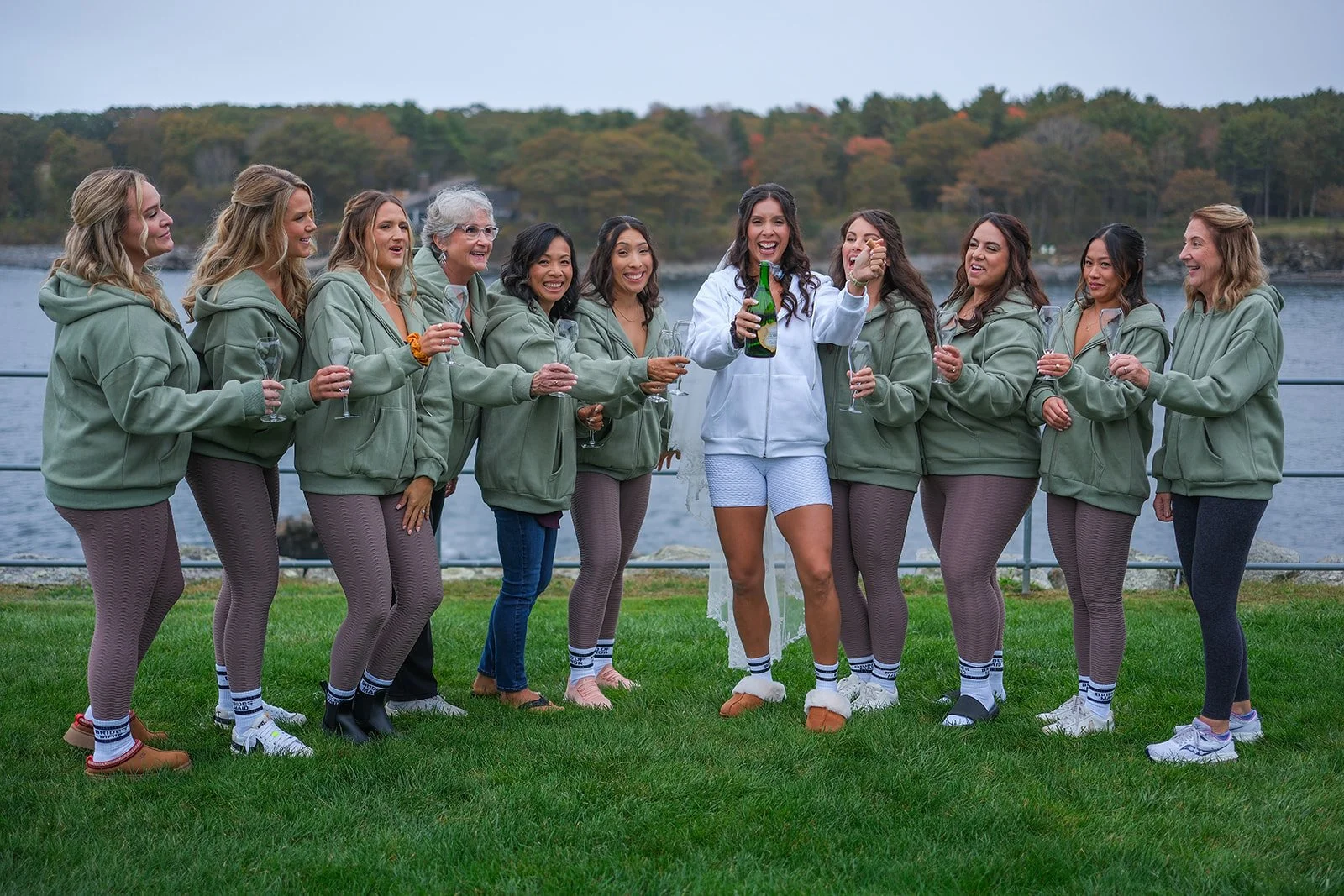 Bride and bridesmaids in matching sage hoodies toasting champagne on ocean lawn during York Maine wedding morning