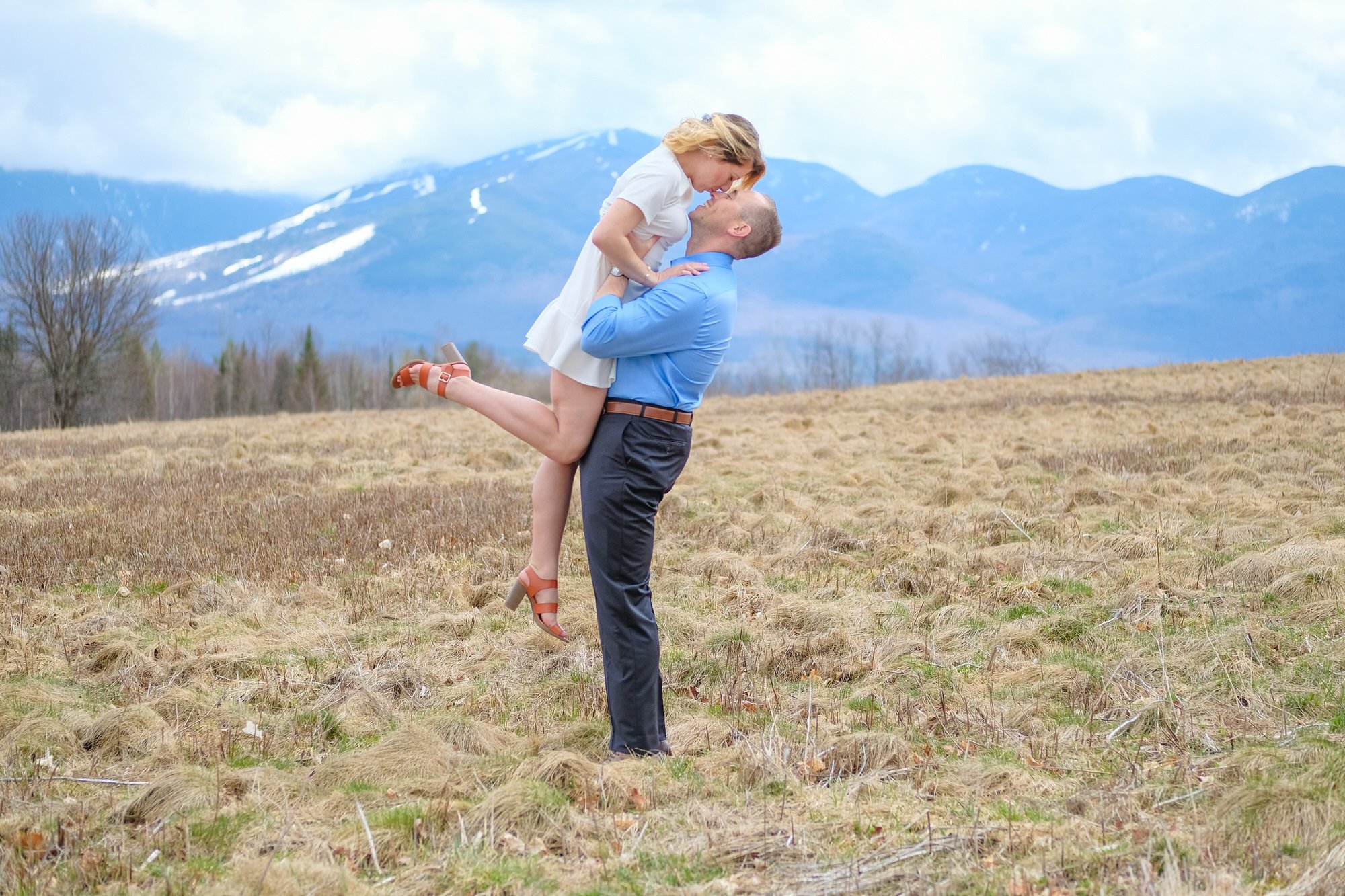 Couple sharing a joyful kiss in an open field with snow-capped White Mountains in the background during their spring elopement in Franconia Notch, NH