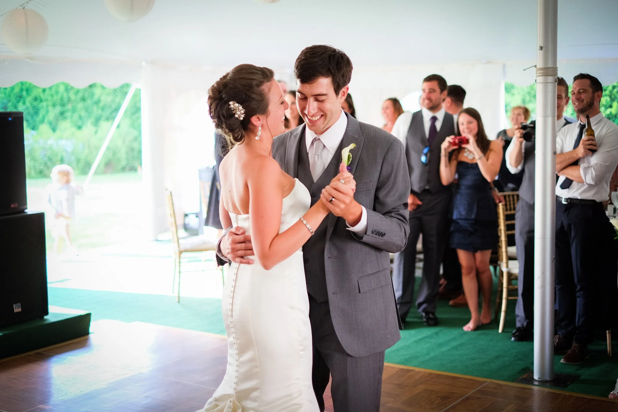 A bride and groom share their first dance at wedding reception with guests watching and taking photos inside a decorated tent.