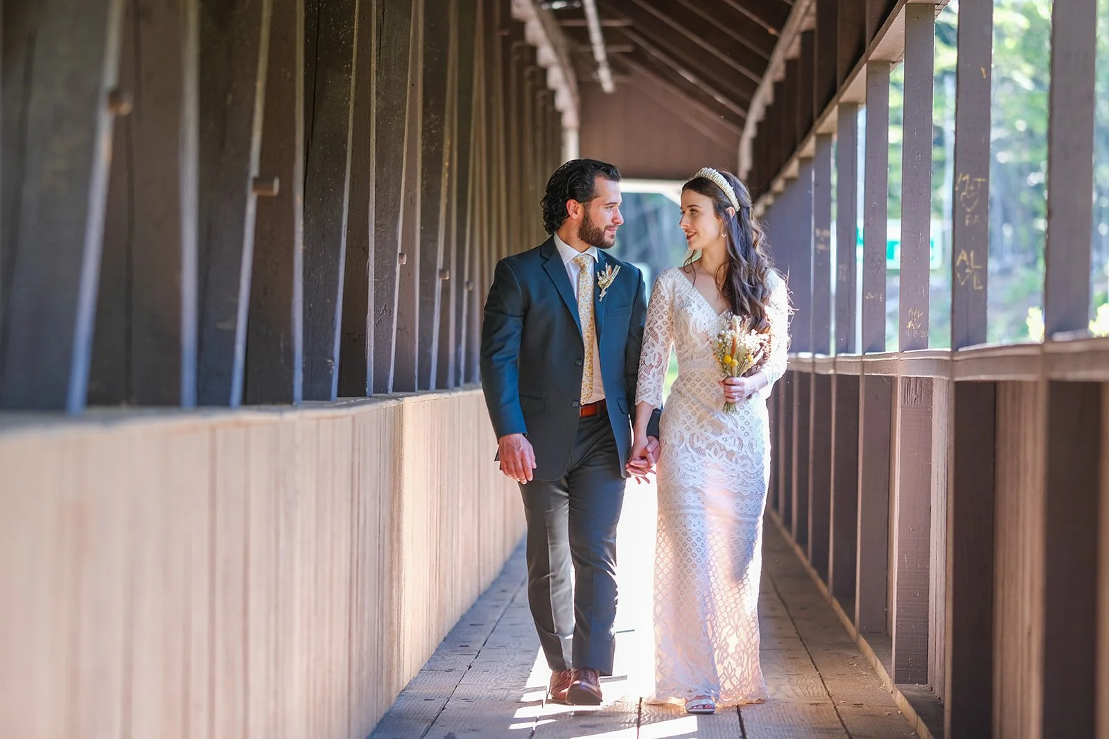 A bride and groom walking hand in hand through a wooden covered bridge during their wedding day. The bride is holding a bouquet and wearing a white lace dress, while the groom is in a dark suit with a patterned tie.
