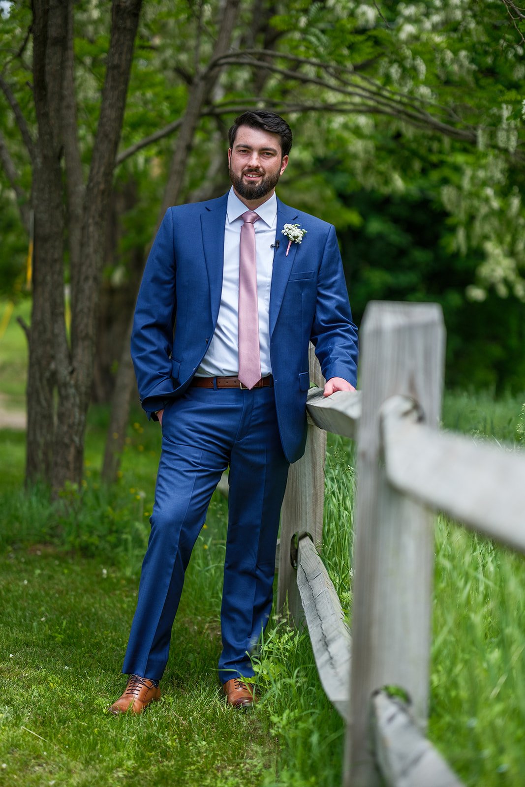 A man in a blue suit with a pink tie and white shirt, standing outdoors beside a wooden fence, surrounded by green trees and grass.