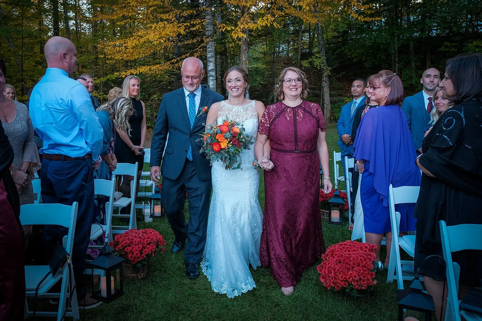 A bride holding a bouquet walks down the aisle with her parents at an outdoor wedding ceremony in a wooded area, surrounded by seated guests and floral decorations.