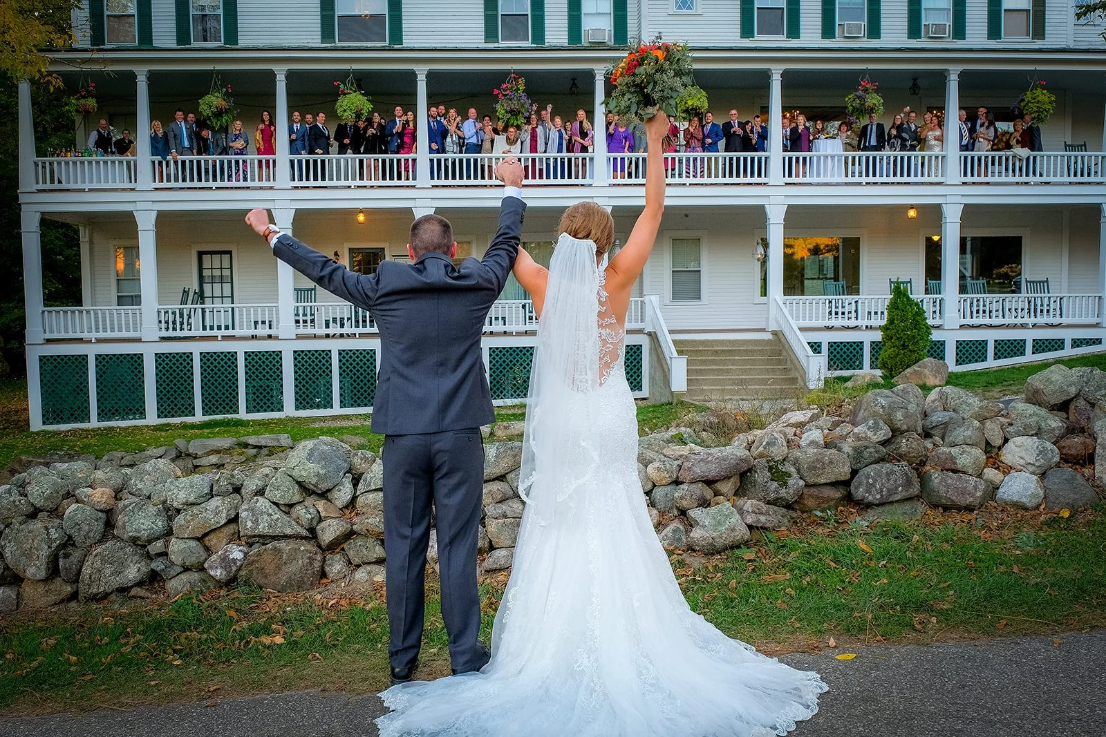 A newlywed couple with raised hands stands in front of a large gathering of guests on a house's porch, celebrating their wedding.