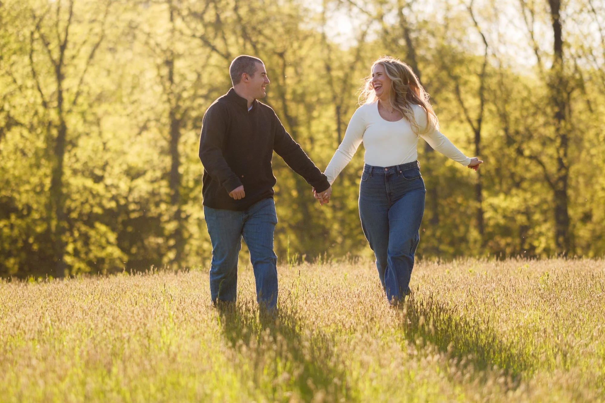 Laughing couple holding hands and walking through a golden sunlit meadow at Maudslay State Park in Newburyport, MA