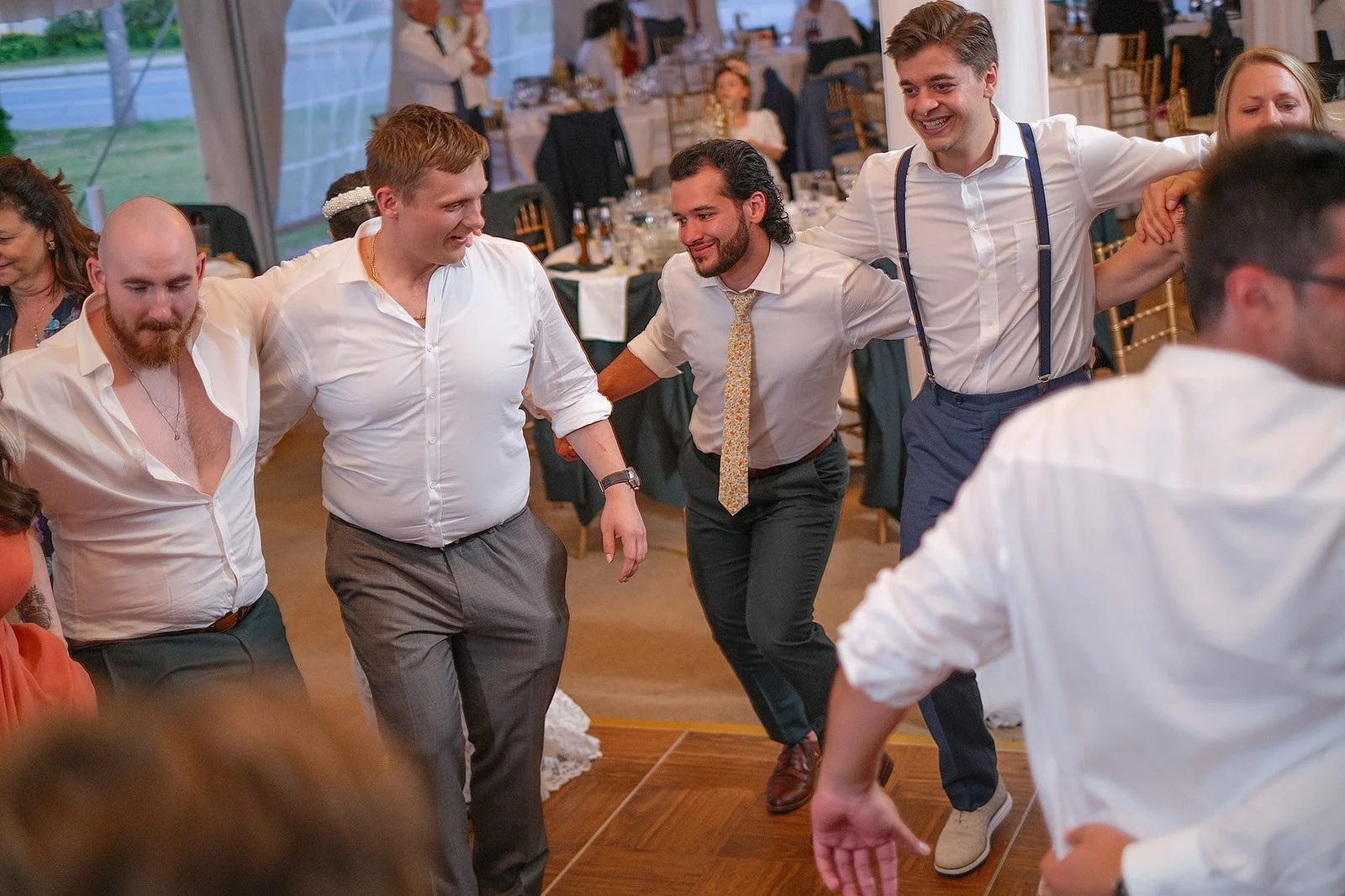 A group of men dancing together at a celebratory event, holding hands and smiling.