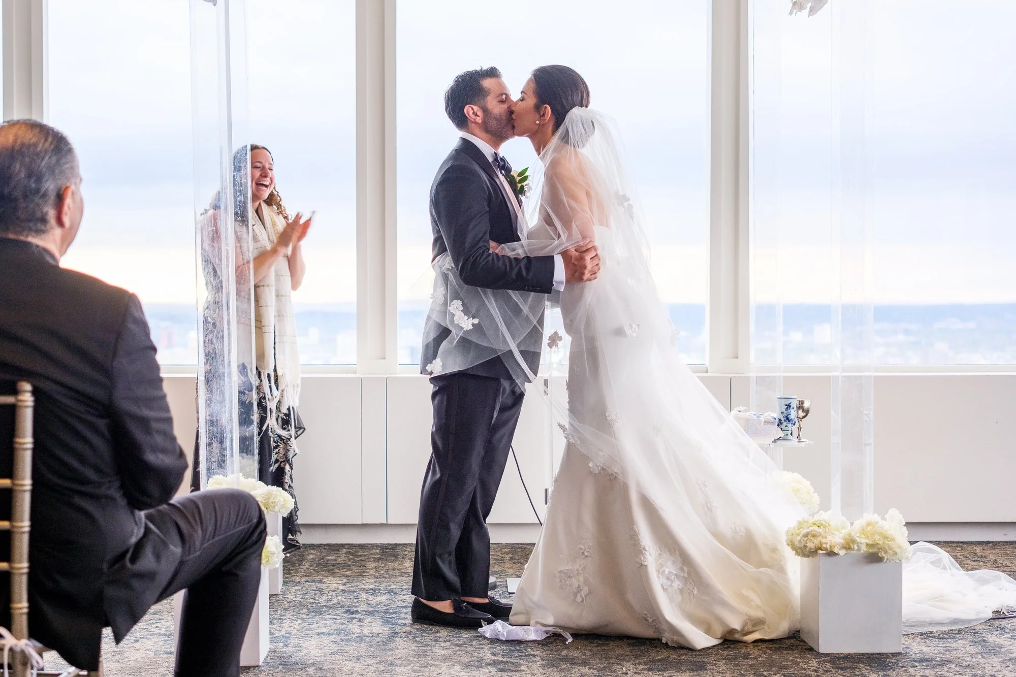 Bride and groom sharing their first kiss as a married couple under the chuppah at the UMass Club Boston as officiant applauds
