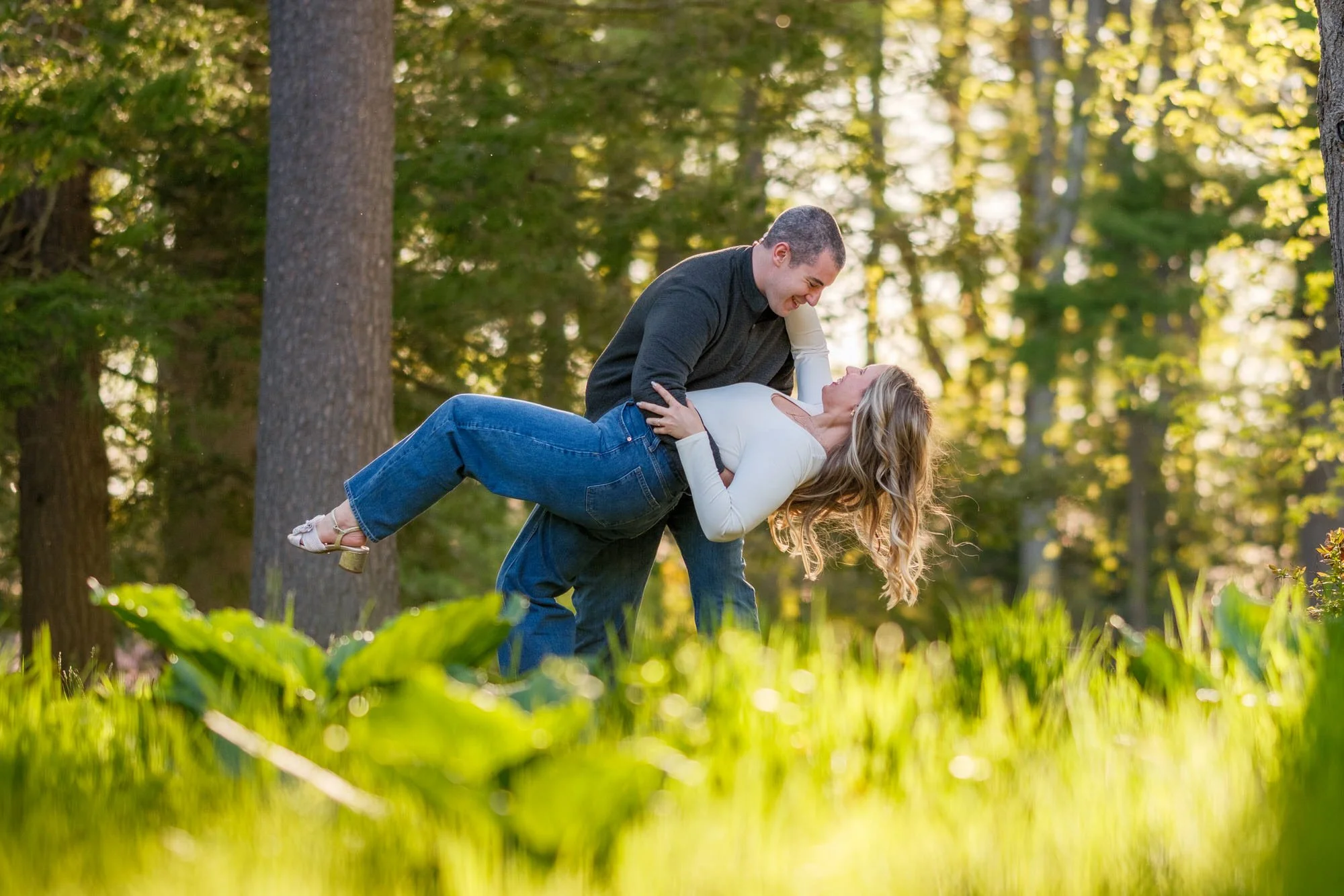 Groom dipping and lifting his laughing fiancée in a sun-drenched pine forest meadow at Maudslay State Park