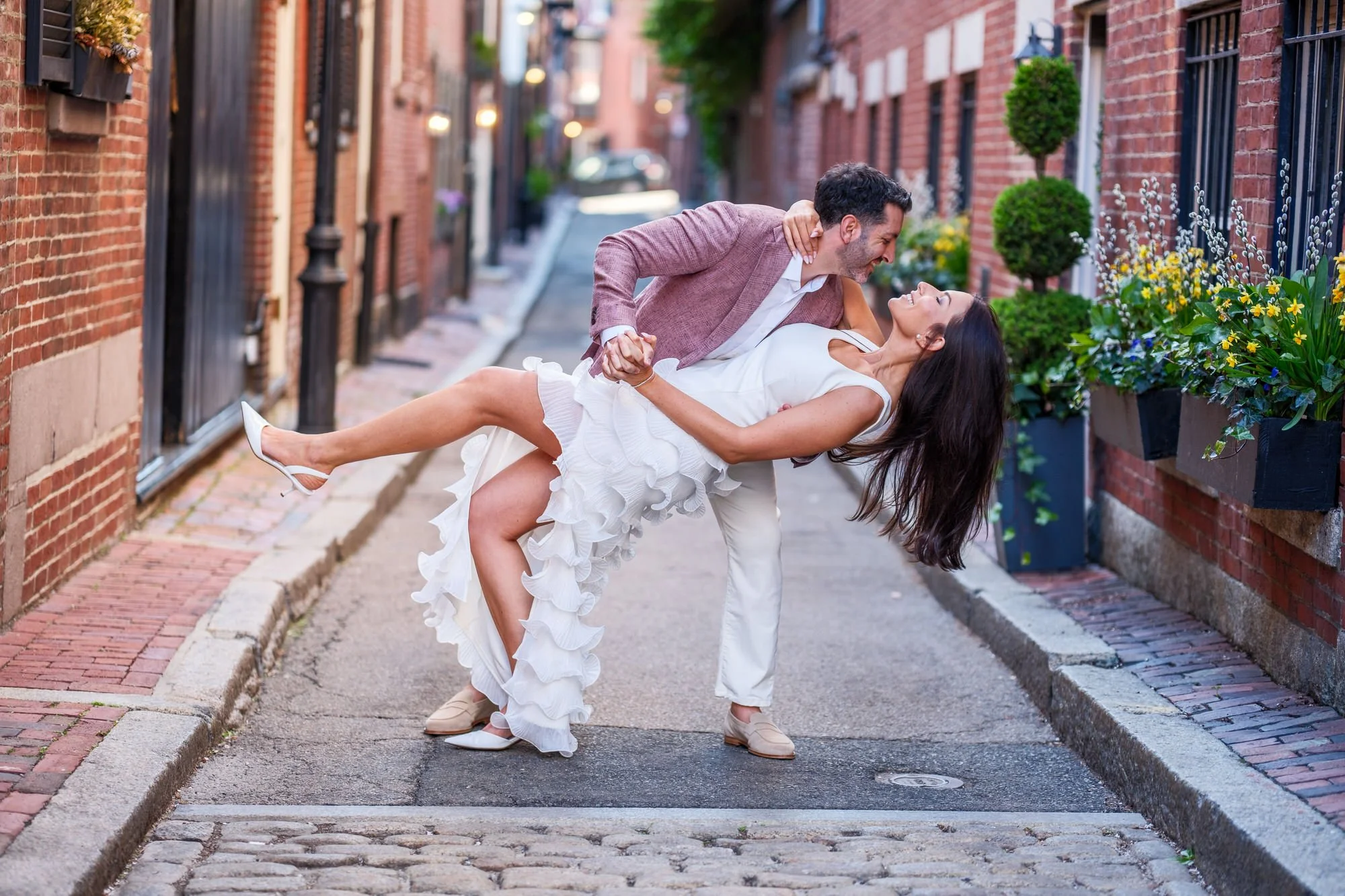 Charlie dipping Lauren dramatically on Acorn Street in Beacon Hill, her white ruffle gown and hair flowing as she laughs, with spring flower boxes and cobblestones framing the joyful moment