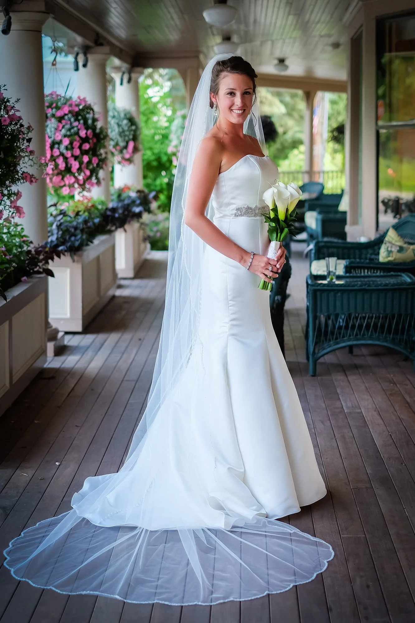 A bride in a white wedding dress holding a bouquet of calla lilies, smiling indoors with flower arrangements and outdoor greenery visible in the background.