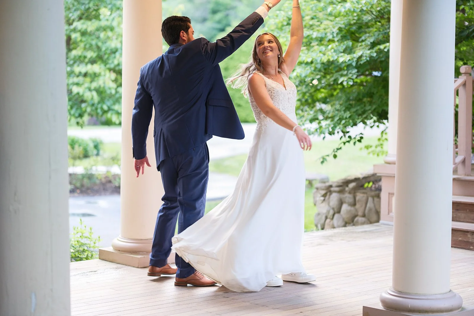 A couple dancing during their wedding outside, with the bride in a white gown and the groom in a dark blue suit, on a porch with white columns and green trees in the background.