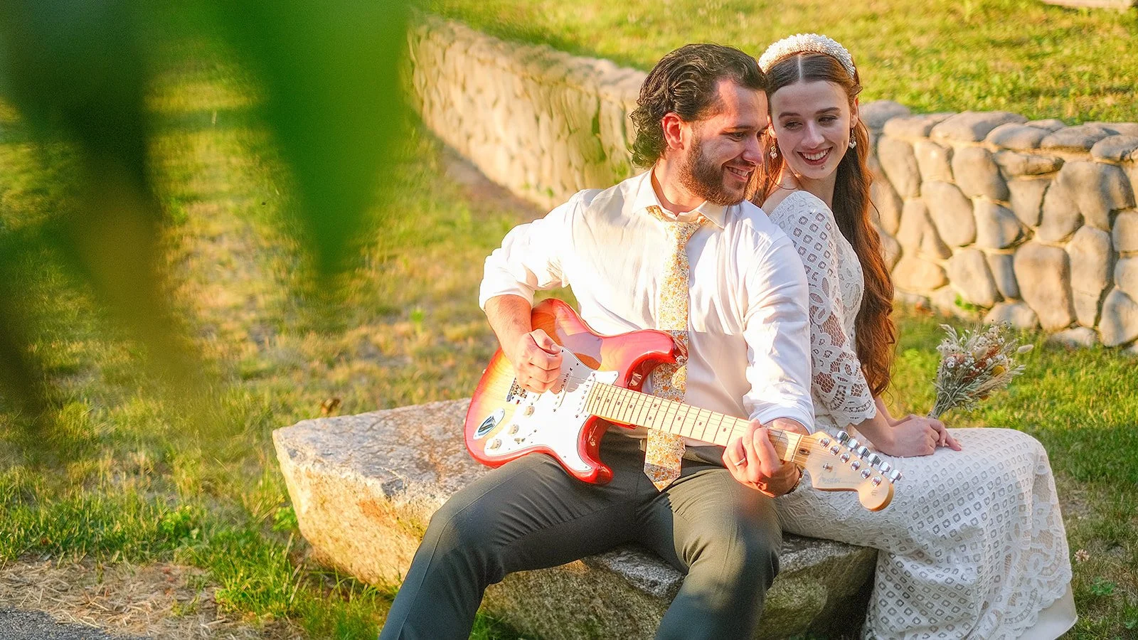 A man and woman sitting on a stone bench outdoors, the man playing an electric guitar and the woman holding a bouquet of flowers, both smiling and enjoying the moment.