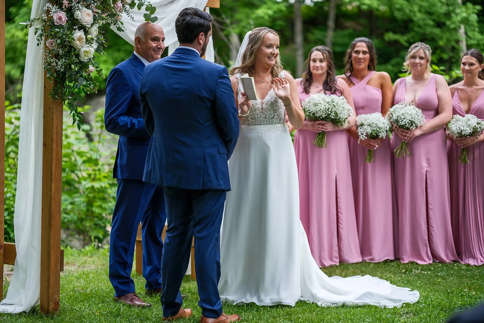 A wedding ceremony taking place outdoors with the bride and groom standing under a floral arch, surrounded by bridesmaids holding bouquets.