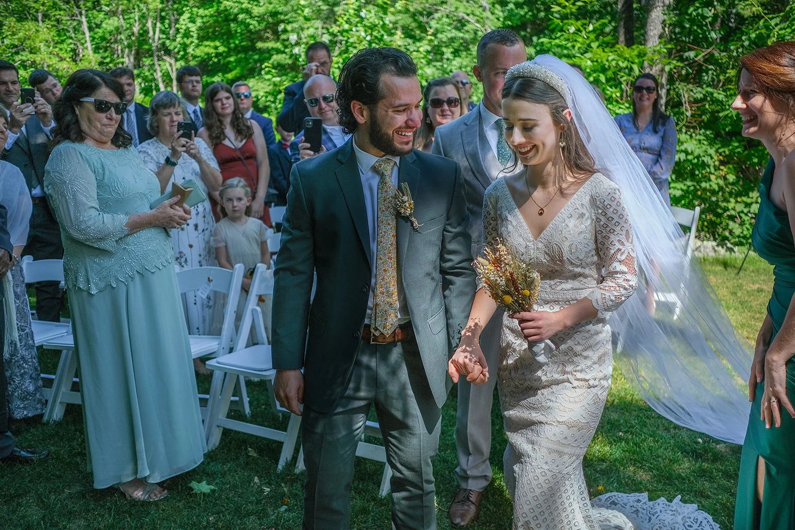 Bride and groom holding hands during outdoor wedding ceremony, surrounded by guests in green outdoor setting.