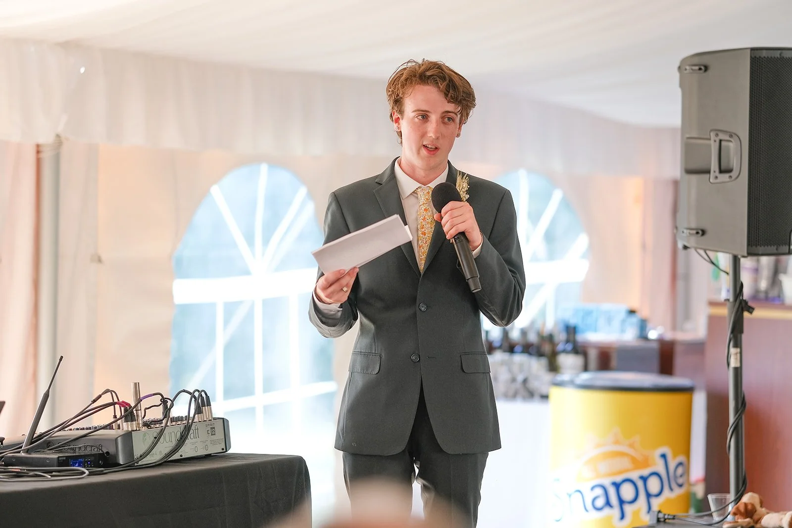 Young man in a grey suit giving a speech at an event, holding a microphone and notes, indoors with a large window and a Snapple cooler in the background.