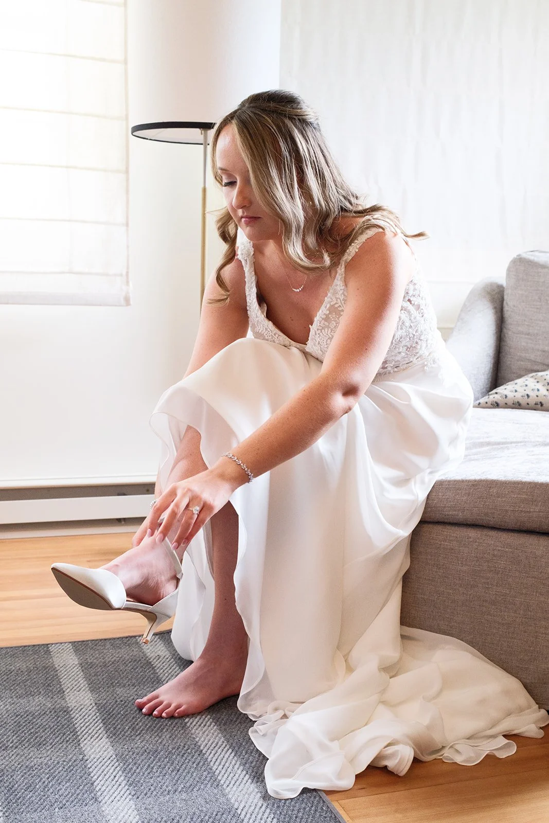 A woman in a wedding dress is sitting on the floor, putting on white high heels in a room with light colored walls and a window.