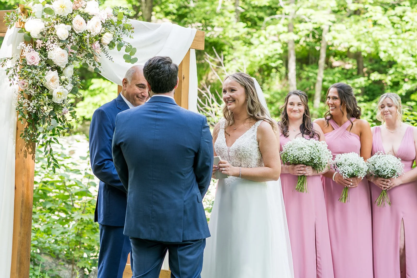 A couple getting married outdoors under a decorated wooden arch with white and pink flowers, with a bride in a white dress and groom in a blue suit, surrounded by bridesmaids in pink dresses holding bouquets, in a lush green forest setting.