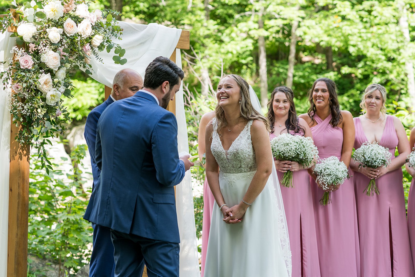 A wedding ceremony outdoors with a bride in a white gown and bridesmaids in pink dresses, standing near a decorated arch with flowers, surrounded by green trees.