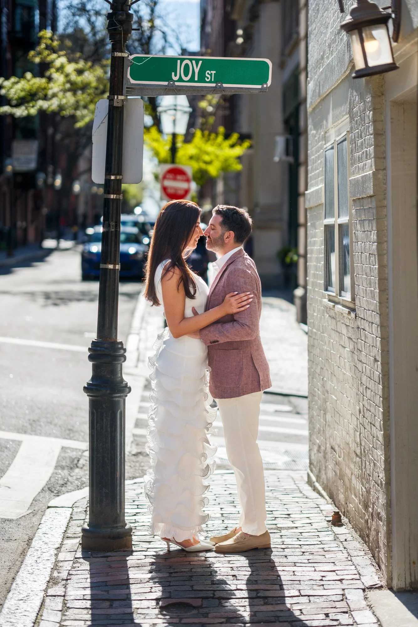 Lauren in her white ruffle gown and Charlie in a blush linen blazer sharing a romantic moment beneath the Joy Street sign on a cobblestone corner in Beacon Hill, Boston