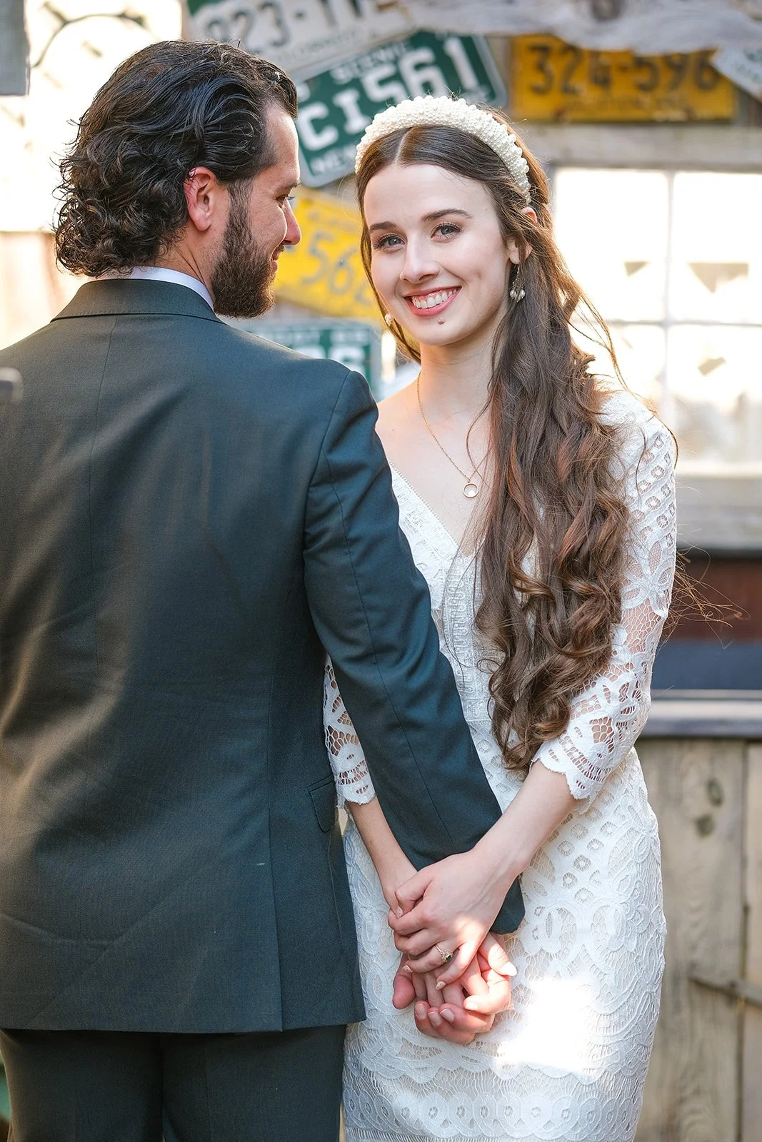 A couple holding hands at their wedding, with the woman smiling and wearing a white lace dress and headband, and the man in a dark suit, standing in front of colorful license plates.