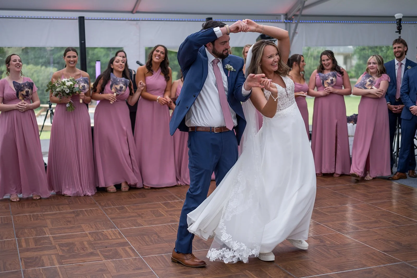 A bride and groom are dancing at their wedding reception, surrounded by bridesmaids in pink dresses and groomsmen in blue suits, under a tent with a wooden dance floor.