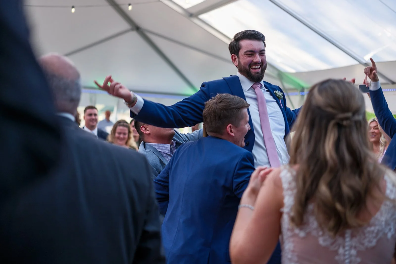 People celebrating at a wedding reception, with a man in a blue suit and pink tie being lifted by others under a tent.