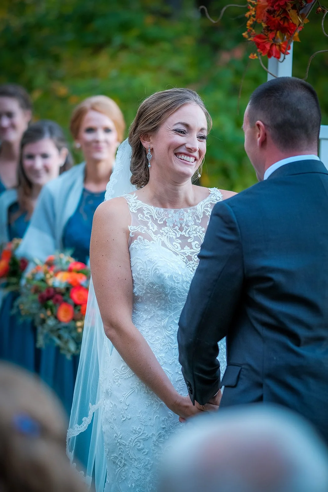 A bride and groom exchange vows outdoors during a wedding ceremony, with bridesmaids and guests watching in the background.