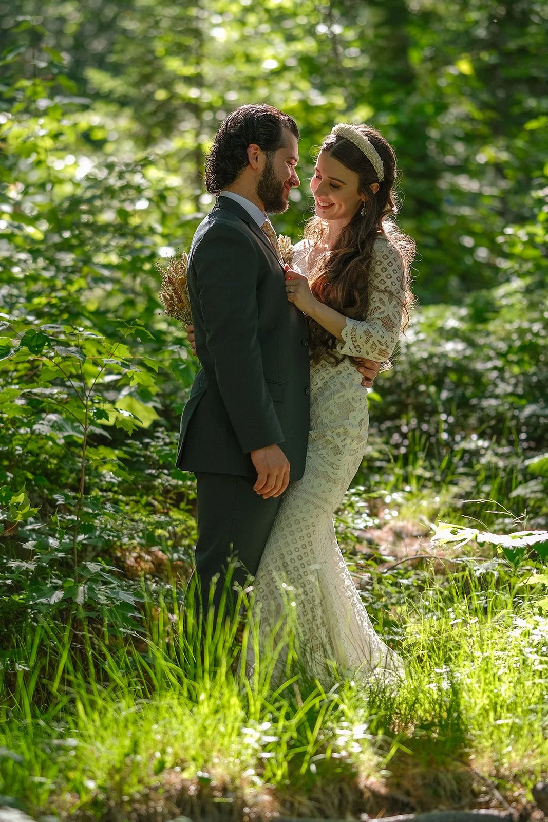 A couple in wedding attire standing close in a lush, sunlit forest with green foliage surrounding them, gazing at each other lovingly.