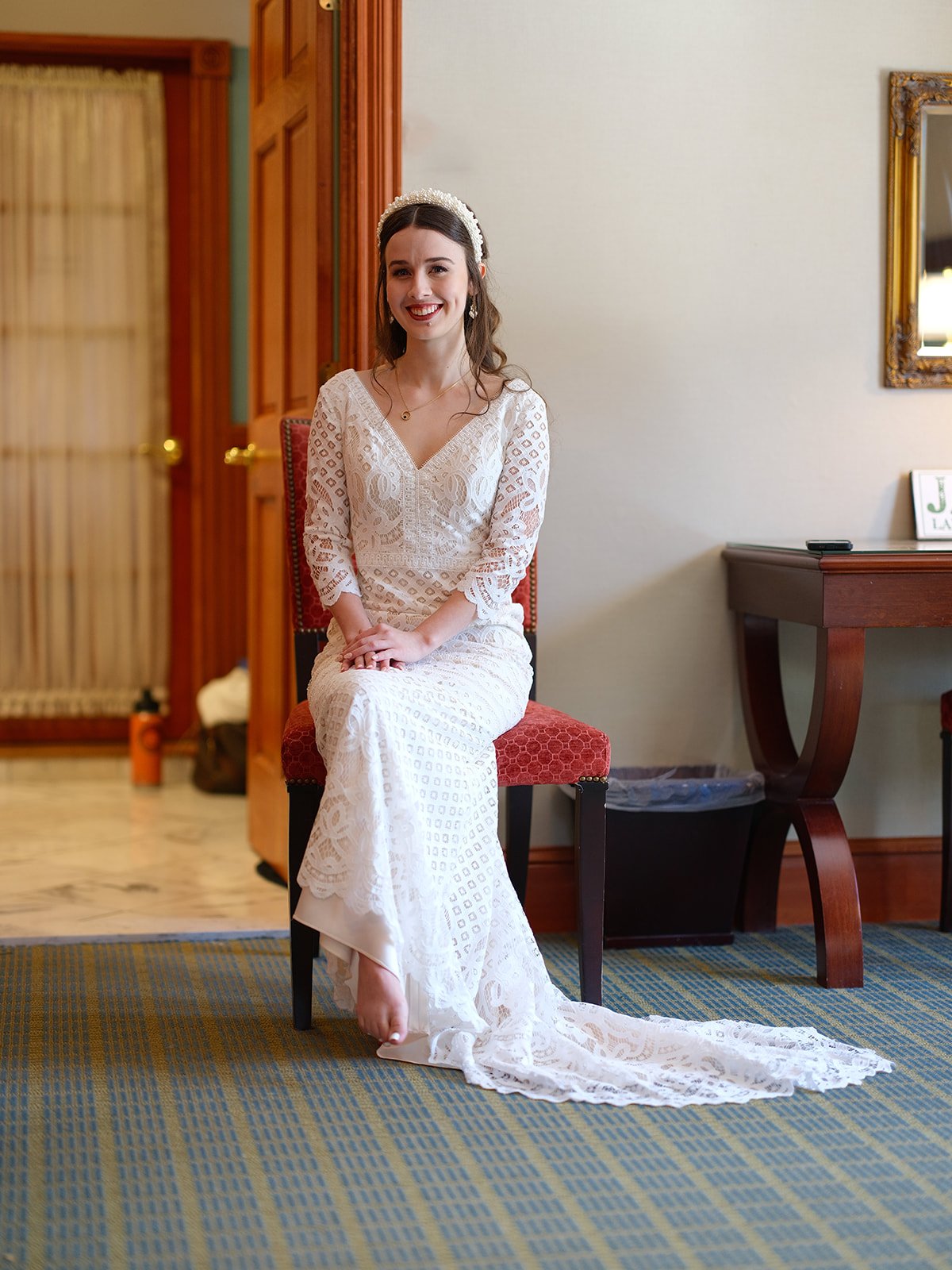 Woman in white lace wedding dress sitting on a red chair, smiling, wearing a headband, in a room with wooden accents and a mirror.