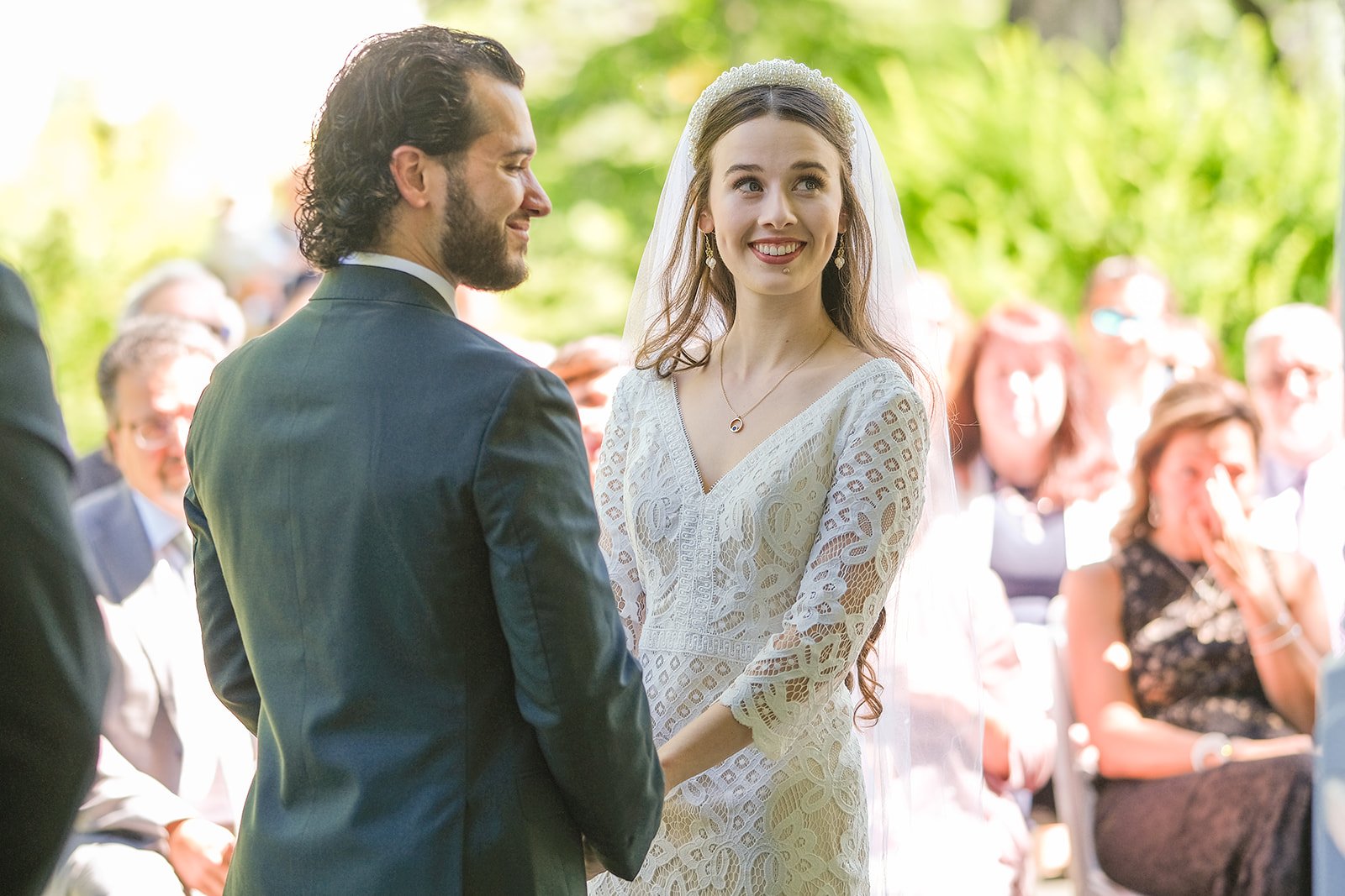 A bride and groom stand facing each other at an outdoor wedding ceremony, surrounded by seated guests, with lush greenery in the background.