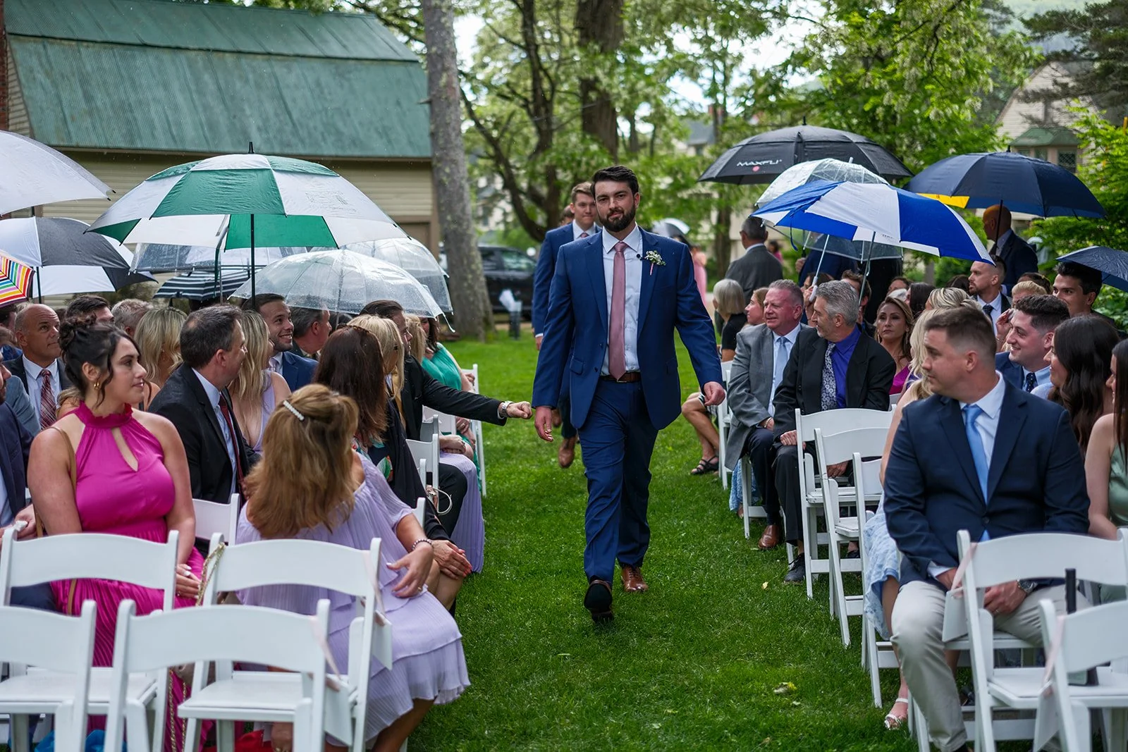 A groom in a blue suit walking down an outdoor wedding aisle between seated guests holding umbrellas due to rain, with lush green trees and buildings in the background.