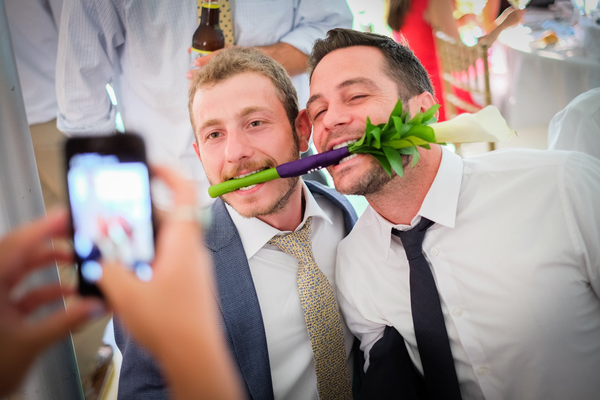 Two men at a celebration taking a photo, one holding a flower in his mouth, smiling while being photographed.